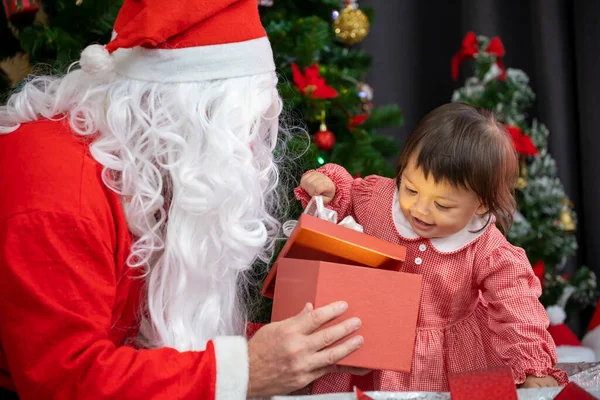 Beyaz bebek Noel Baba 'dan hediye alıyor. Noel kutlaması için süslenmiş Noel ağacının yanında.