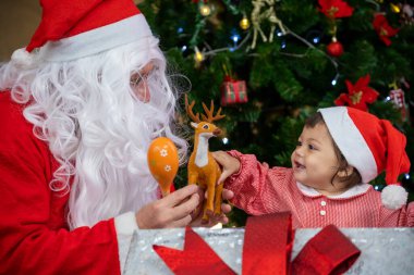 Beyaz bebek Noel Baba 'dan hediye alıyor. Noel kutlaması için süslenmiş Noel ağacının yanında.