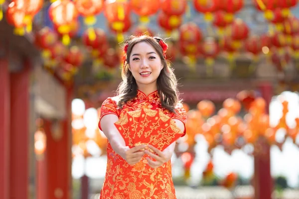 Asian woman in red cheongsam qipao dress is holding red envelope for money saying \'May you have great luck and profit\' inside Chinese Buddhist temple during lunar new year for best wish blessing