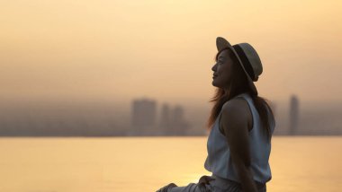 Silhouette of pretty tourist woman is relaxing and enjoying her rooftop swimming pool view with cityscape at sunset background for vacation and travel