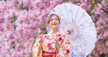 Geleneksel kimono elbiseli Japon kadın elinde şemsiyeyle ve tatlı Hanami Dango tatlısıyla bahar Sakura Festivali 'nde kiraz çiçeği ağacının yanında fotokopi alanı ile yürürken.