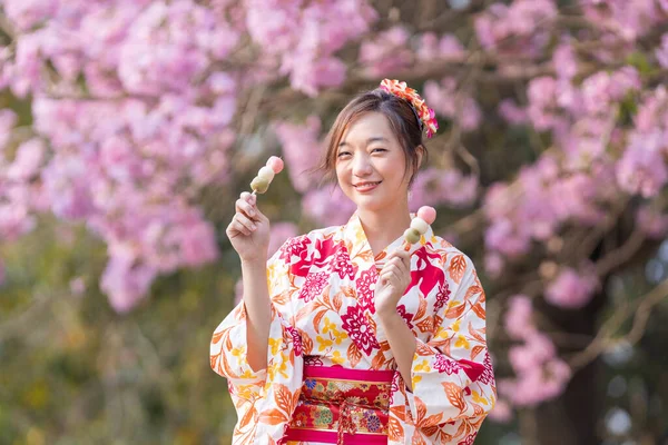 春の桜まつりの公園を散策しながら 甘い花見団子のデザートを手にした着物姿の日本人女性 — ストック写真