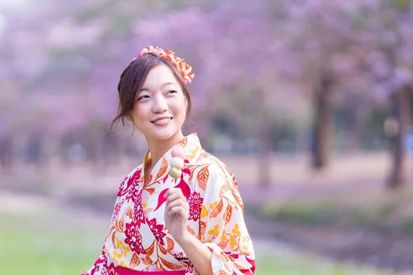 春の桜まつりでは 桜の木の公園を歩いている間に甘い花見団子デザートを持っている伝統的な着物を着た日本人女性コピースペース — ストック写真