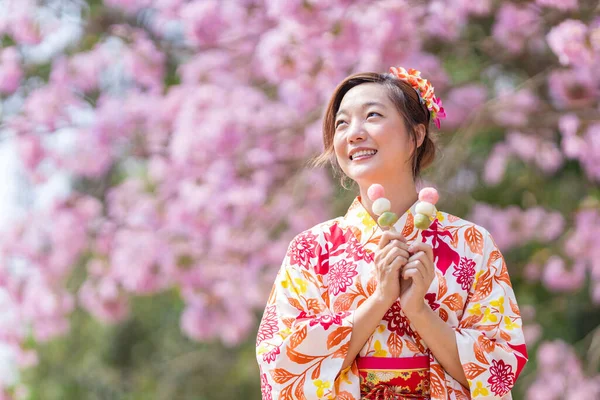 春の桜まつりの期間中 桜の木の公園を歩きながら甘い花見団子デザートを持つ伝統的な着物姿の日本人女性 — ストック写真