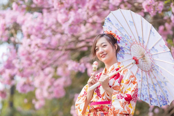 春の桜まつりの期間中 桜の木の公園を歩いている間 傘と甘い花見団子デザートを持つ伝統的な着物姿の日本人女性 — ストック写真
