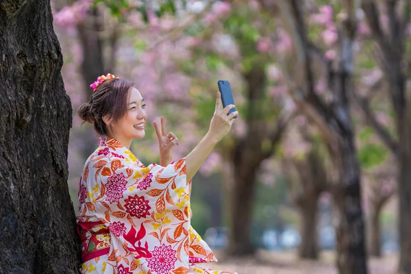 stock image Japanese woman in traditional kimono taking selfie with peace gesture while walking in the park at cherry blossom tree during spring sakura festival usage