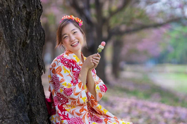 春の桜まつりの期間中 桜の木の公園を歩いている間 甘い花見団子デザートを持つ伝統的な着物姿の日本人女性 — ストック写真