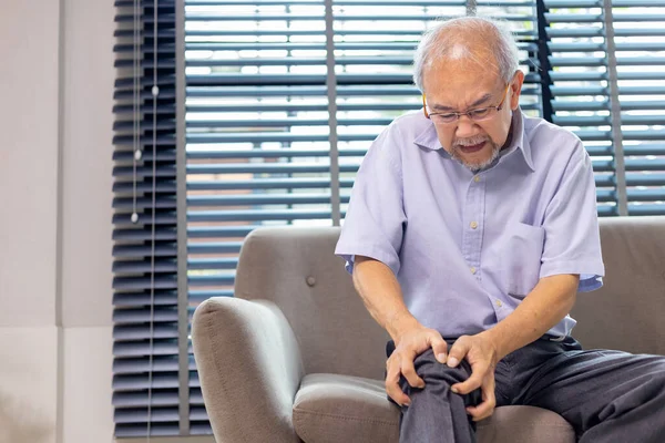 患有膝关节骨关节炎症状的亚裔老人坐在家里的沙发上抱怨着 带着供手术和理疗用的复印空间 — 图库照片