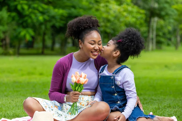 African mother holding bouquet of tulip flowers and daughter kissing each other while happily enjoying picnic in the public park during summer for family love and care in mother\'s day celebration