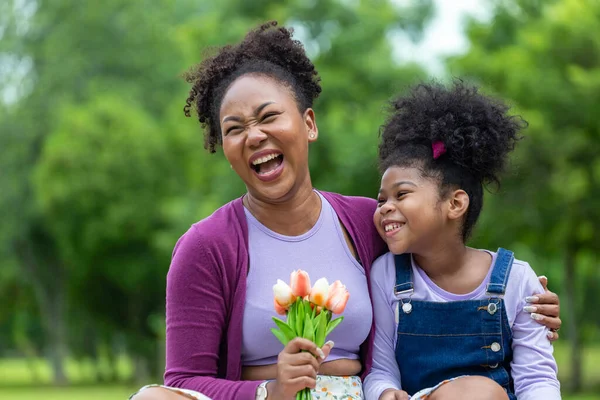 African mother is holding bouquet of tulip flowers and the daughter hugging each other while happily enjoying picnic in the public park during summer for family love and care in mother\'s day celebration