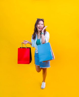 Yazın moda olan güzel Asyalı kadın, reklam ve tanıtım etkinliği konsepti için sarı arka planda izole edilmiş indirimli satış için mutluluk içinde gülümsüyor ve alışveriş çantası tutuyor.