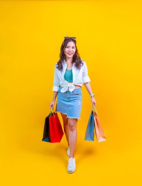 Yazın moda olan güzel Asyalı kadın, reklam ve tanıtım etkinliği konsepti için sarı arka planda izole edilmiş indirimli satış için mutluluk içinde gülümsüyor ve alışveriş çantası tutuyor.