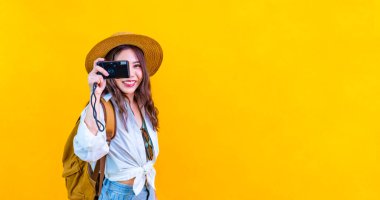 Modayı takip eden güzel Asyalı kadın yolcu, seyahat etmek için kamera kullanıyor ve sarı arka planda izole edilmiş yaz tatili ve fotokopi alanı kullanıyor..