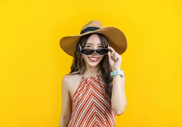 Portrait studio shot of Asian woman in sexy dress with straw hat and sunglass smiling in happiness isolated on yellow background for travel and summer vacation