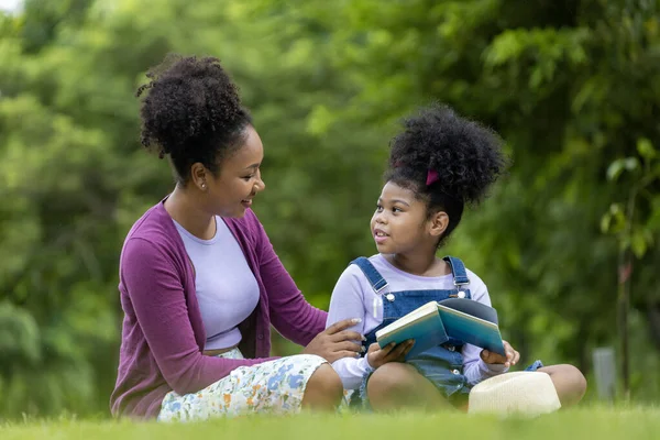 アフリカ系アメリカ人の母親は 教育と幸福の概念のための公園で夏のピクニックをしている間に読むように彼女の若い娘を教えています — ストック写真