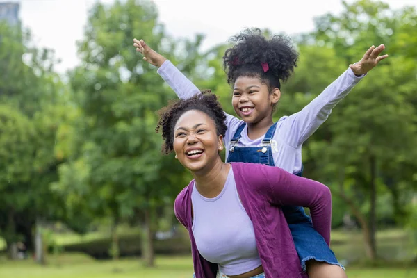 アフリカ系アメリカ人の母親は 幸福と幸福のために公園で夏のピクニックをしながら 彼女の若い娘と一緒にピギーバックに乗って遊んでいます — ストック写真