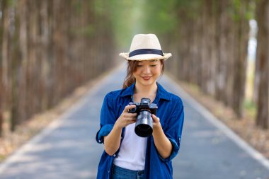 Asyalı turist kadın, seyahat ve fotoğrafçılık için ağaç sütunuyla yolda yürürken profesyonel kamerayla milli parkta fotoğraf çekiyor.