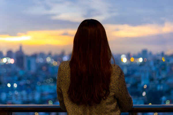 Woman looking and enjoying sunset view from balcony with sun setting behind skyscraper in busy urban downtown with loneliness for solitude, loneliness and dreaming of freedom lifestyle