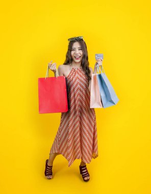 Yazın moda olan güzel Asyalı kadın gülümsüyor ve alışveriş çantası tutuyor. İndirim için kredi kartı, reklam ve tanıtım etkinliği için sarı arka planda izole edilmiş.