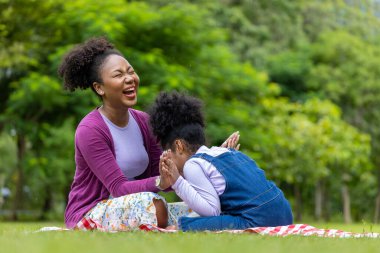 African American mother is laughing while playing patty cake with her young daughter while having a summer picnic in the public park for wellbeing and happiness