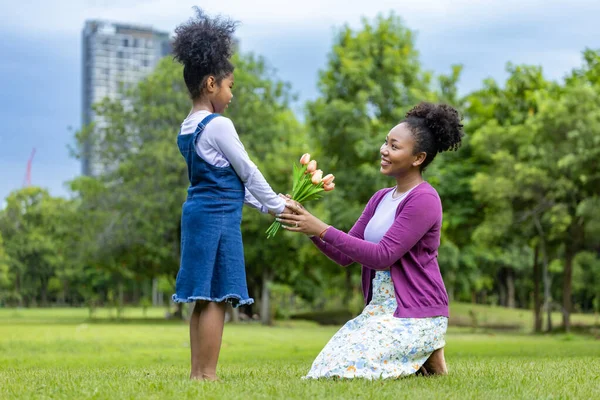 アフリカの娘は 母の日に家族の愛とケアのための夏の間に公園でピクニックを楽しみながら 母のためのチューリップの花の花束で彼女の母親を驚かせます — ストック写真