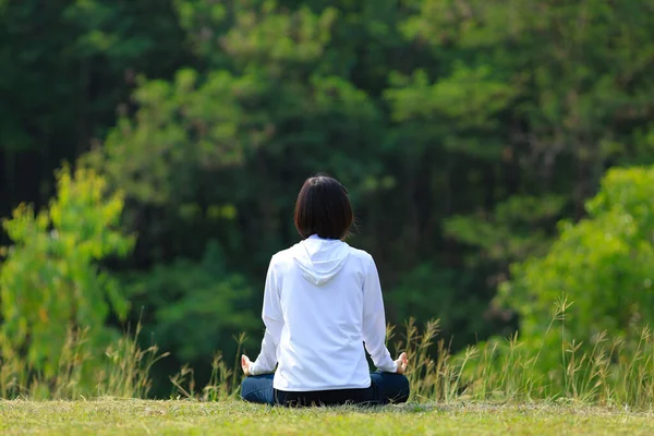 Kapüşonlu bir kadının arka görünüşü, yaz aylarında çim çayırlarında rahat bir şekilde meditasyon yogası yapıyor. Sağlıklı bir ruh ve zihin için sabah ışığı ile iç huzuru bilgeliğinden mutluluk elde etmek için.