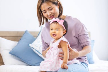 Asyalı anne güzel gülümseyen kızını kucağında tutarken aile mutluluğu ve ebeveynlik kavramı için yatakta kaliteli zaman geçiriyor.