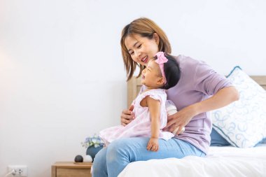 Asyalı anne güzel gülümseyen kızını kucağında tutarken aile mutluluğu ve ebeveynlik kavramı için yatakta kaliteli zaman geçiriyor.