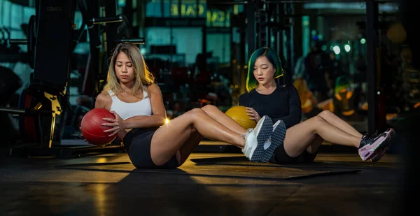 两名亚洲女子体操伙伴进行坐姿斜扭运动 将重量药球放在地板上 以增强腹肌和骨骼肌 — 图库照片