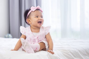 Tatlı pembe elbiseli Asyalı bebek gülümseyerek sağlıklı çocuk ve sevimli kız portresi kullanımı için mutlulukla yatakta oturuyor.