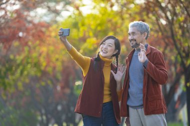 Mutlu Asyalı kız ve kıdemli baba sonbahar boyunca halk parkında akçaağaç ve ginkgo ağacıyla yürürken sonbahar gezileri ve aile mutluluğu için selfie çekiyorlar.