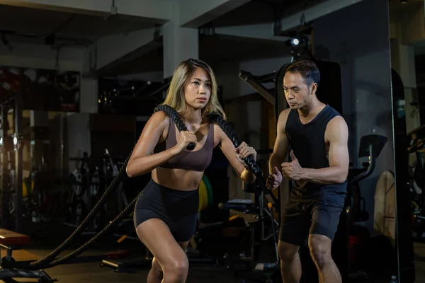 亚洲妇女通过拉绳进行了强壮有力的核心体重训练 她的辅助教练鼓励在体育馆内进行高级锻炼和锻炼 — 图库照片
