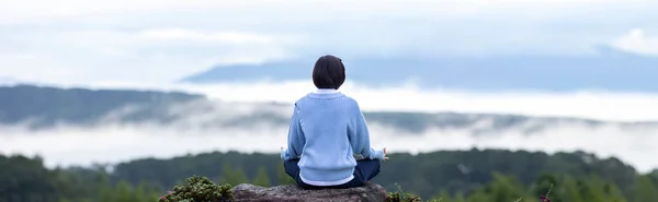 Panorama back view of woman in hoodie is relaxingly practicing meditation yoga at top of mountain with mist and fog in summer to attain happiness from inner peace wisdom for healthy mind and soul