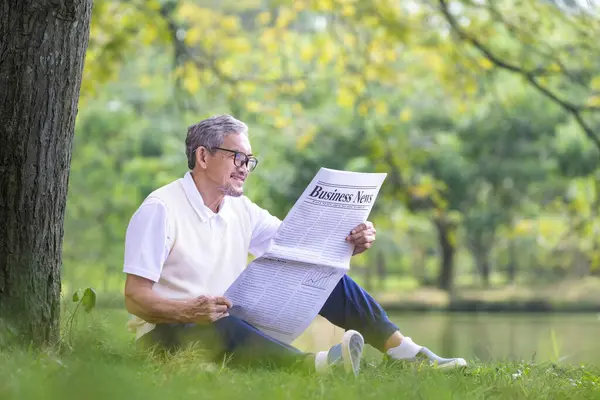 Senior Asian man reading a business newspaper while sitting under the tree by the lake at public park for recreation, leisure and relaxation in nature