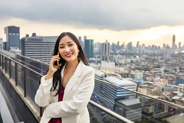 Asian business woman in formal suit is phone calling customer while standing outside skyscraper building for marketing, connection, corporate work, real estate, housing and urban development