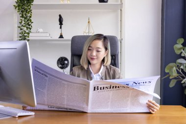 Ofiste çalışan Asyalı iş kadını, iş çalışanları için günlük resmi kıyafetlerle sosyal eğilimi güncellemek için gazete okurken bilgisayar kullanıyor.