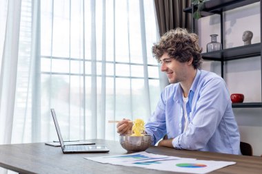 Beyaz adam, hazır erişte yerken ve serbest ve girişimci işleri için istatistik tablosunu incelerken ev ofisinde çalışıyor.