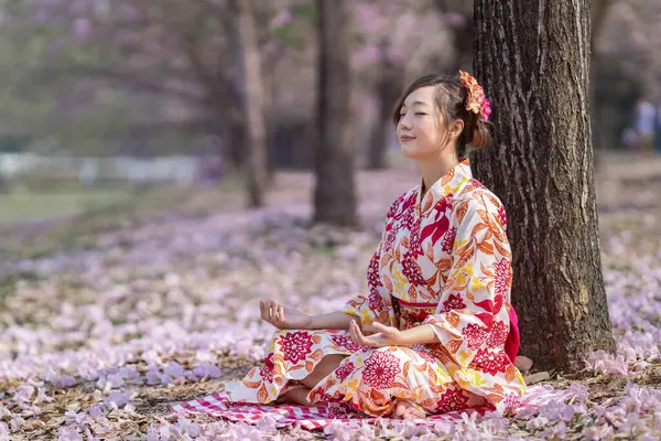 Kimono elbiseli Japon kadın kiraz çiçekleri açarken Kiraz ağacı altında meditasyon yapıyor. İç huzur, farkındalık ve zen çalışması için.