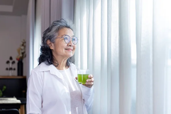 Senior Asian lady drinking glass of herbal supplement drink while looking out the window with copy space for nutrition boosting and healthy organics product consumption concept
