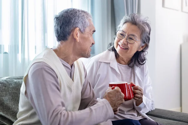 Couple of healthy Asian senior father and mother sitting on couch with happy smile at retirement home drinking hot tea to celebrate their holiday together for elder care to spending valuable time