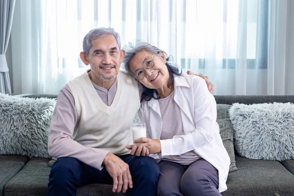 Couple of healthy marriage Asian senior father and mother sitting together with glass of milk and happy smile on cozy couch in retirement home during winter for elder care to spending precious time