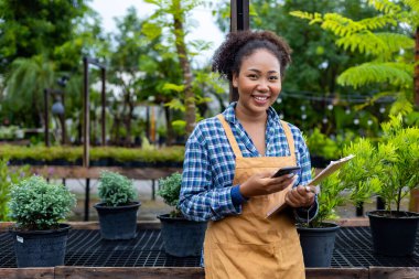 Afrikalı Amerikalı bahçıvan Nad fidanlık sahibi kozalaklı ağaç yetiştirme bahçesinde çalışırken elinde bir pano tutuyor.