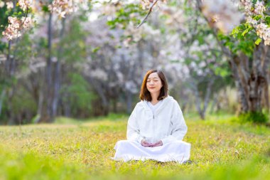 Asyalı kadın, iç huzur, farkındalık ve zen uygulama konsepti için kiraz çiçeklerinin açtığı mevsimde çiçek ağacının altında meditasyon yapıyor.