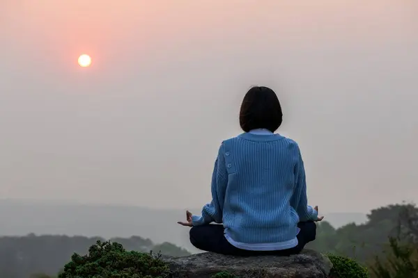 Back view of woman is relaxingly practicing meditation yoga mudra on mountain top with rising sun during summer to attain happiness from inner peace wisdom for healthy mind and soul