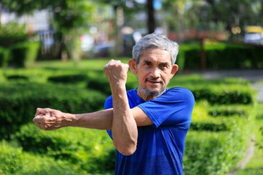 Kıdemli Asyalı adam ısınma egzersizleri sırasında kolunu ve omuz kaslarını esnetiyor. Sabahleyin halk parkında sağlıklı ve uzun ömürlü olmak için egzersiz yapıyor.