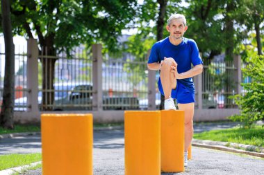 Kıdemli Asyalı adam ısınma egzersizi sırasında bacak kaslarını esnetiyor. Sabahleyin halk parkında sağlıklı ve uzun ömürlü olmak için egzersiz yapıyor.