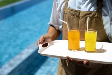 Garson yaz boyunca tatil ve seyahat konsepti için lüks oteldeki yüzme havuzunun yanındaki müşteriye iki bardak buzlu tropikal meyve suyu taşıyor.