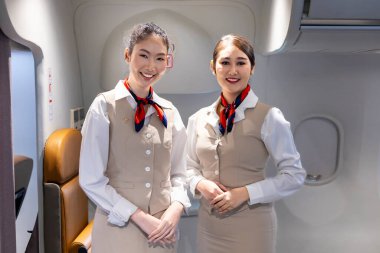 Asyalı uçuş görevlisi ekibi, arka planda koltuğu olan yolcuyu karşılamak için koridorun ortasında gülümseyerek poz veriyor.