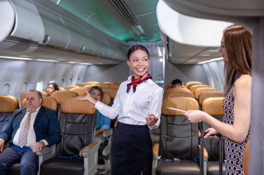 Uçuş görevlisi birinci sınıf yolcuları karşılıyor ve yolcu biletini kontrol ediyor ve uçak ve havayolu ulaşımı için koltuğunun yerini gösteriyor.
