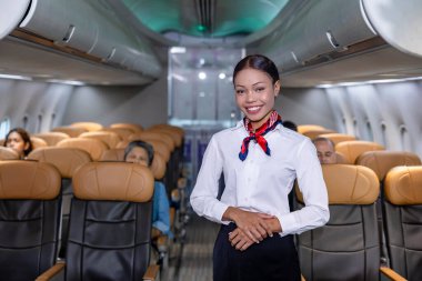 Arka planda koltuğu olan yolcuyu karşılamak için koridorun ortasında gülümseyen yüzüyle poz veren Afrikalı uçuş görevlisi.